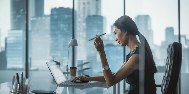 A woman sits at a desk in a modern office with large windows, working on a laptop and holding a pen while looking at the screen thoughtfully. City skyscrapers are visible in the background.