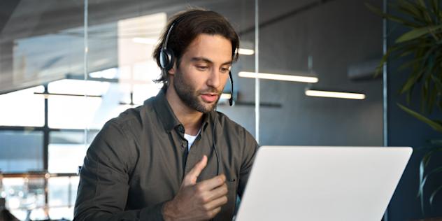 A man wearing a headset sits at a desk in front of a laptop, speaking and gesturing with his hand during a video call in a modern office setting.